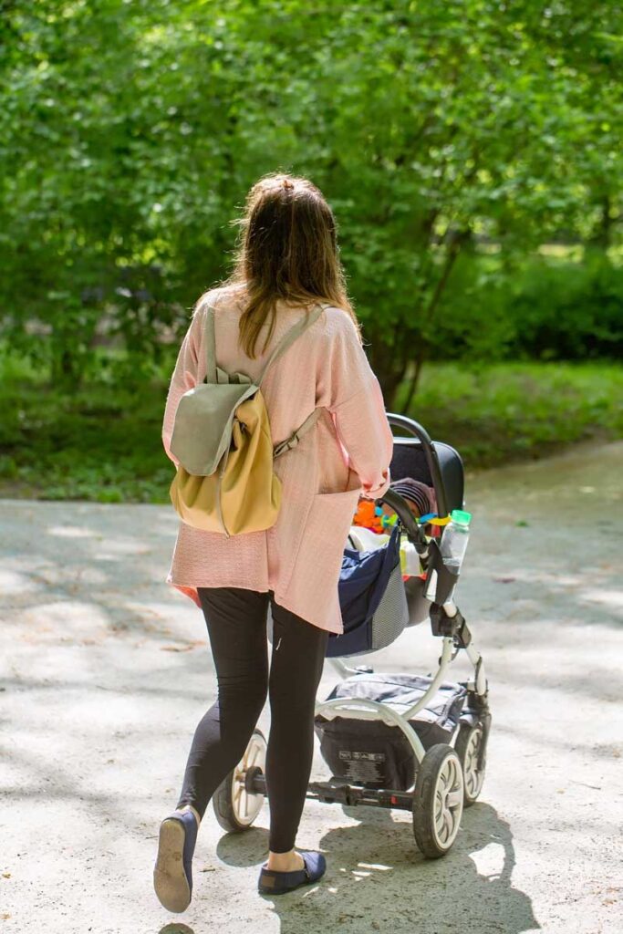 a woman pushes a baby stroller in the park with a few accessories clipped to her stroller.