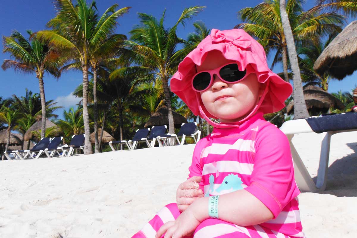 Toddler in sunglasses and pink hat sitting on beach