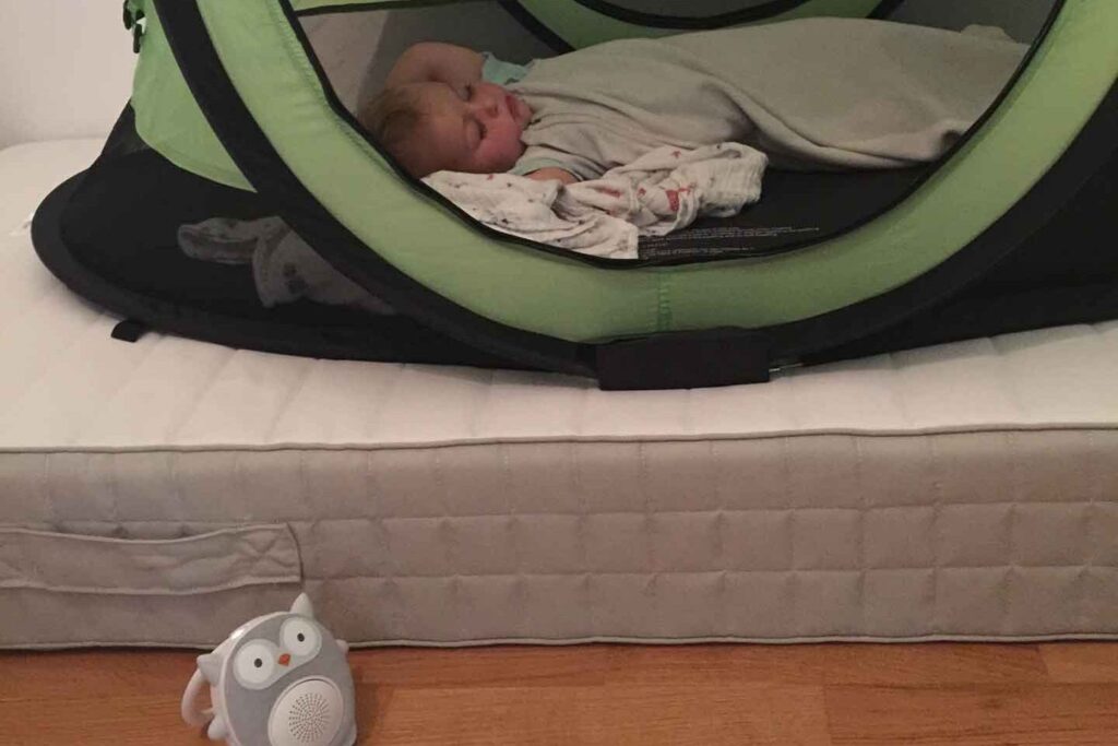 A toddler from BabyCanTravel.com sleeps in a PeaPod Plus travel bed while using a SoundBub white noise machine