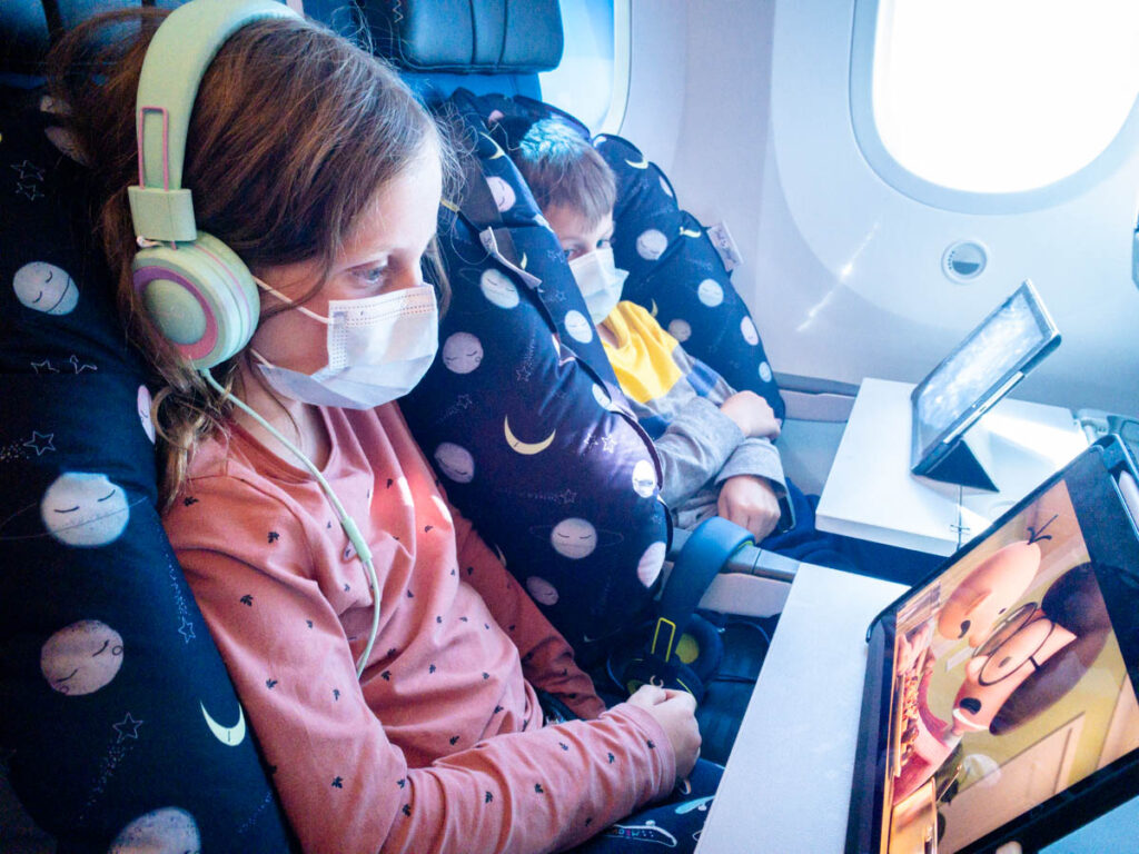 Two kids from the Baby Can Travel family watch movies on their iPads while relaxing into their Seat to Sleep Travel Nest Kids Plane Pillow.