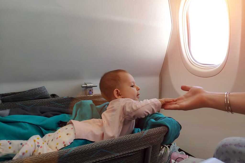 Baby holding parents hand while laying in baby bassinet on airplane.