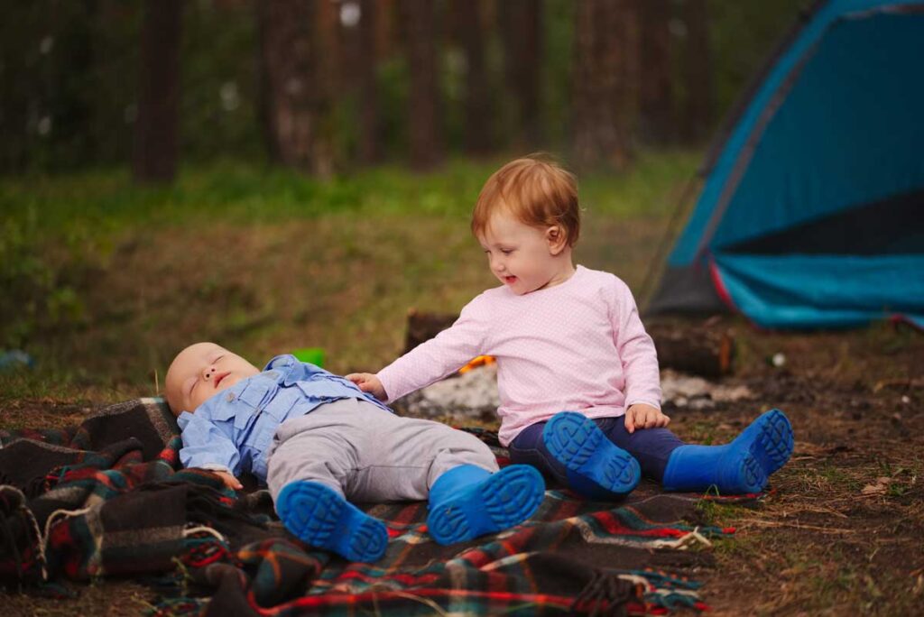 two babies on a family camping trip a sit on a blanket blanket near a tent.