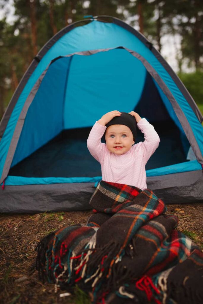 a baby sitting outside a camping tent with a hat and blanket.