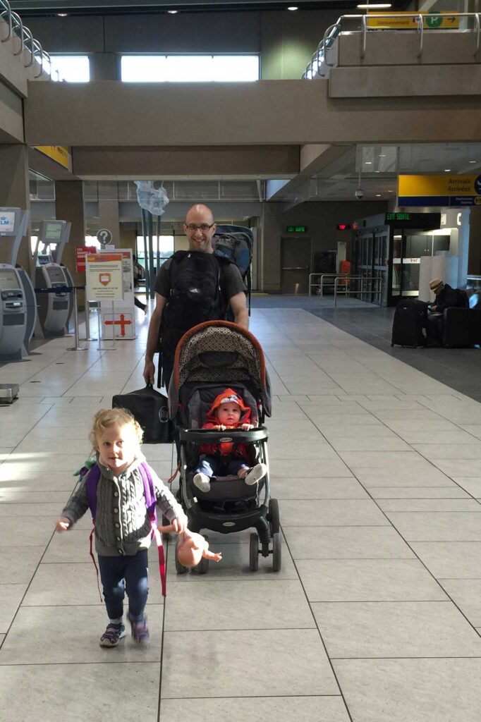 Dan Brewer, of the Baby Can Travel blog, carries a backpack, a pack n play while pushing a baby in a stroller. His toddler runs ahead holding her favorite doll.