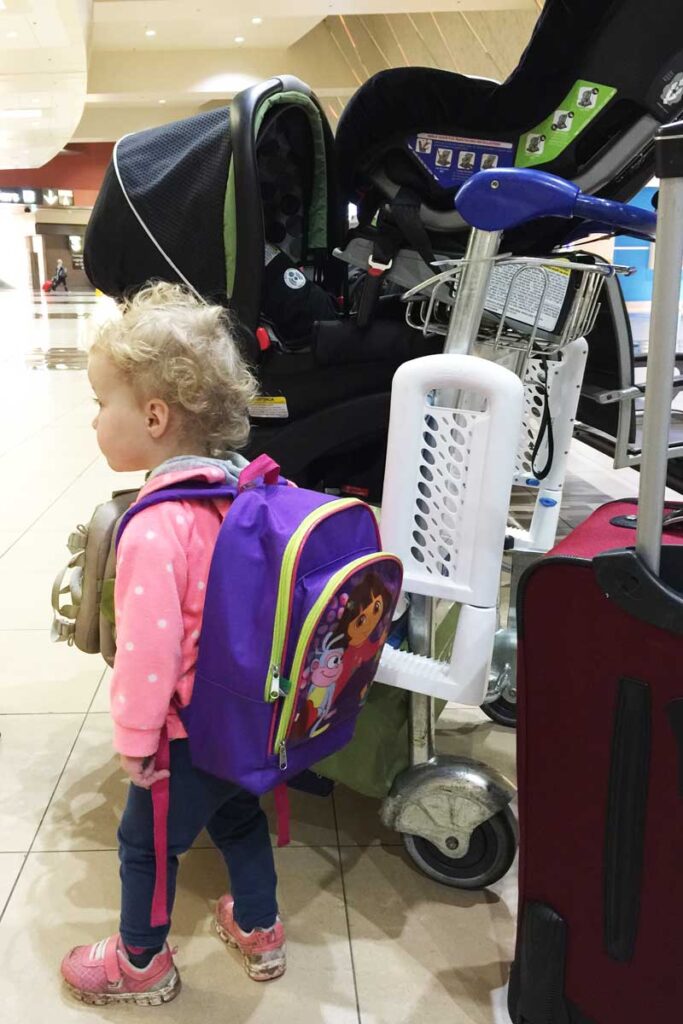 a toddler stands in front of an airport cart filled with baby equipment.