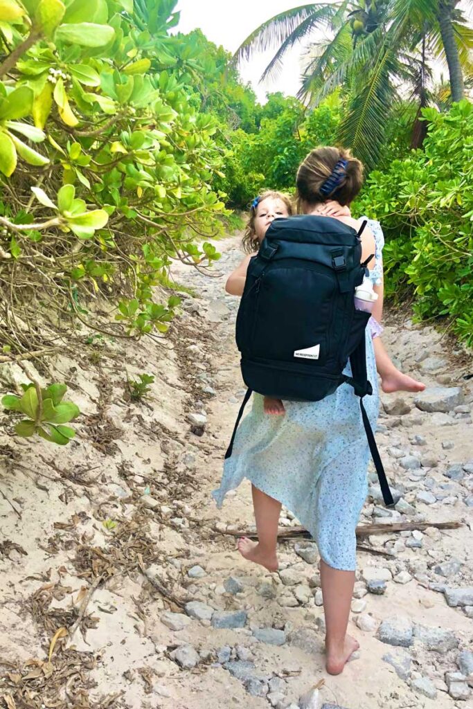 Mother carrying toddler on beach path while wearing black travel backpack diaper bag