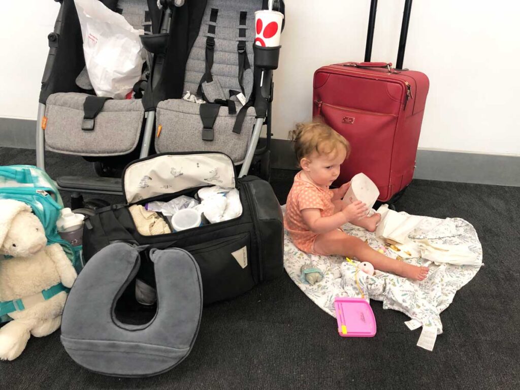 baby sitting on airport floor surrounded by luggage, double travel stroller and open diaper backpack by No Reception Club