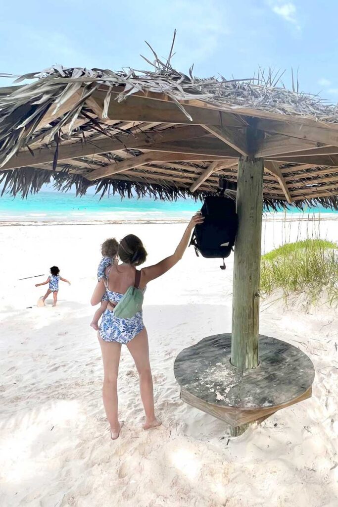 woman holding baby on the beach under a large umbrella with the NRC diaper backpack hanging on umbrella