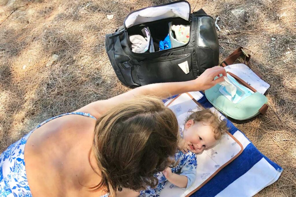 mother changing baby's diaper on the beach, while demonstrating easy access to diaper changing essentials with a diaper backpack and wipes access of the Sidekick by No Reception Club nearby