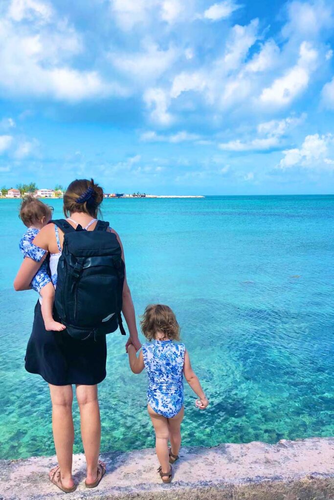 Mother wearing black Getaway bag by No Reception Club holding toddlers hand while carrying baby in front of the turquoise colored ocean