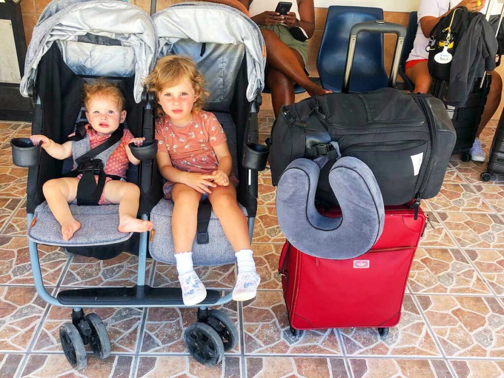 Baby and toddler in double travel stroller with red suitcase holding the No Reception Club bag for travel beside the stroller