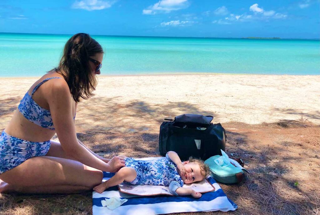 Mother changing baby's diaper on the beach, while demonstrating easy access to diaper changing essential using No Reception Club backpack diaper bag