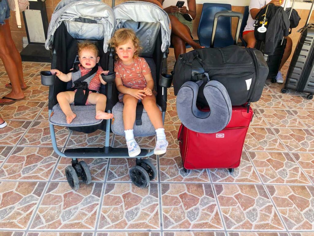 baby and toddler in a grey Zoe Twin Double Stroller at airport next to red suitcase