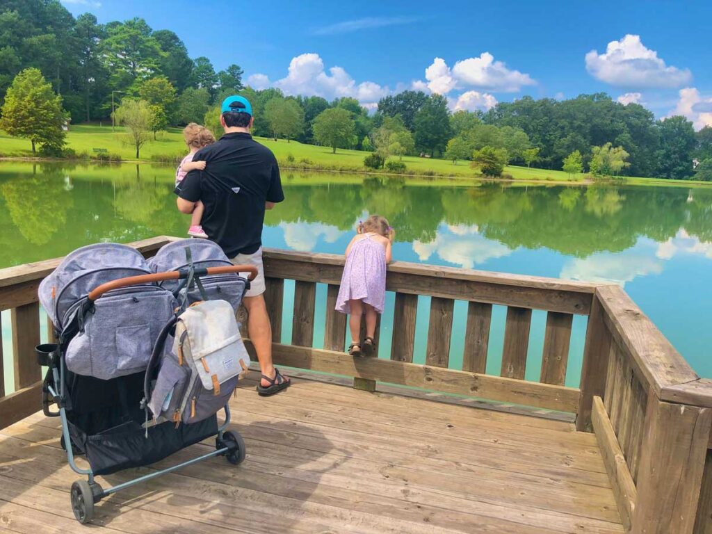 Zoe double stroller on wooden viewpoint with father holding baby and toddler standing in front of it overlooking water