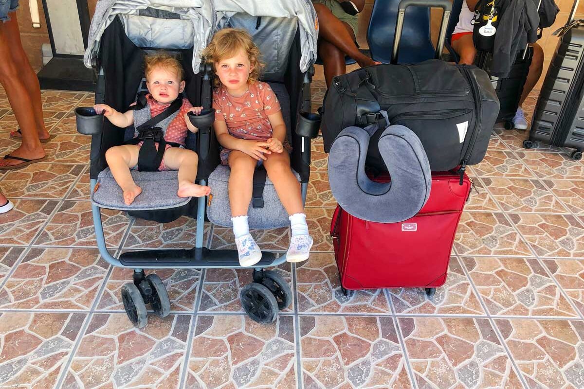 baby and toddler in a grey Zoe Twin Double Stroller at airport next to red suitcase