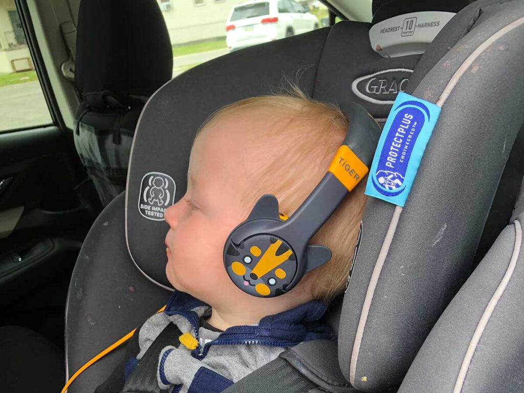 a small child listens to Kidrox Tiger-Ear Toddler Sized Headphones in a car seat.