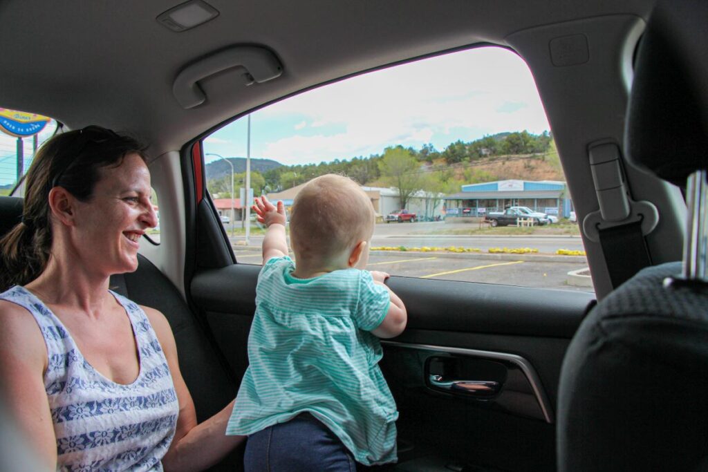 A mother sits in the back seat of a car with her playful 1-year old.