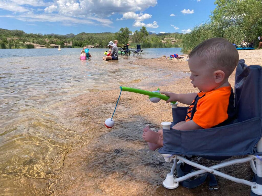 Toddler pretending to fish while sitting in a booster chair in the water. 