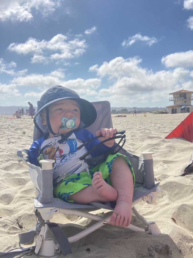 a baby wearing a sun hat sits in a hiccapop OmniBoost Booster Seat on the beach.