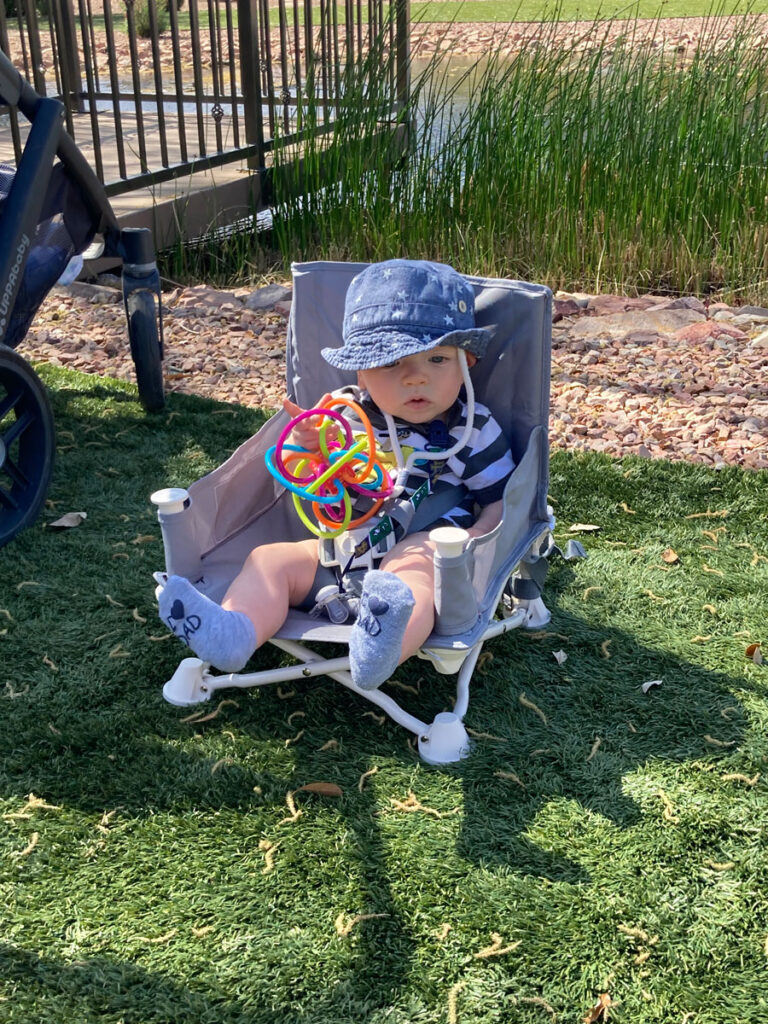 Baby sitting in a booster chair in the grass eating. 