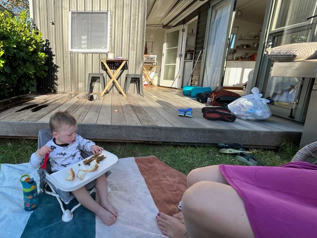 Baby sitting in booster chair that is on top of a blanket eating food, lady in the background. 