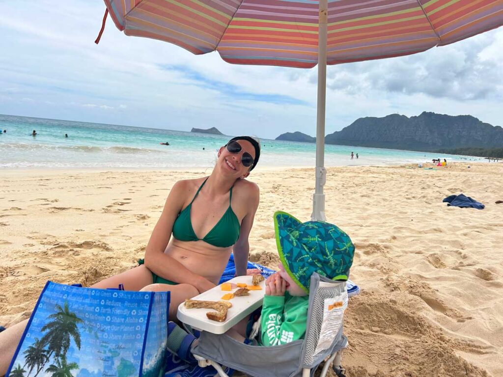 Baby in a hiccapop travel booster chair that is on the beach, with a woman in a green bikini sitting nearby smiling.