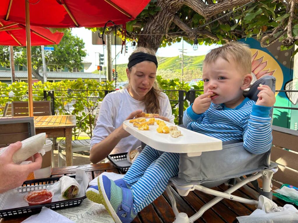 Baby eating food while sitting in booster seat with lady in the background.