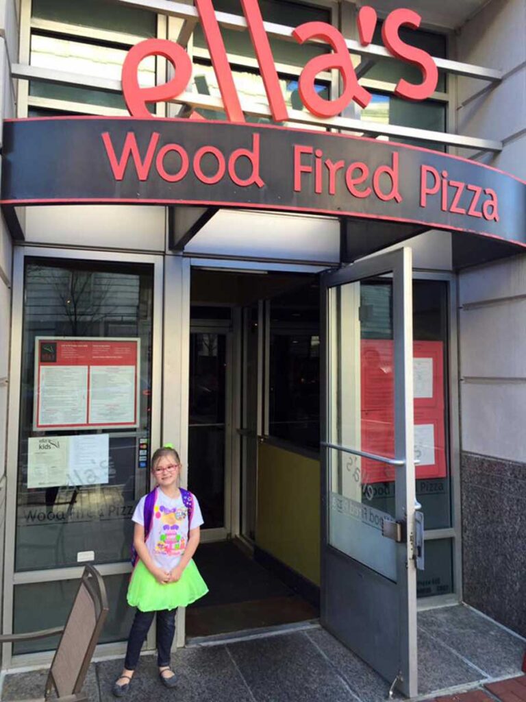 Young girl standing outside ellas pizza restaurant.
