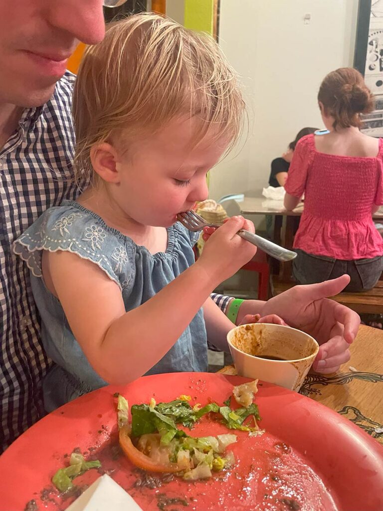 Toddler sitting on man's lap and eating dinner.