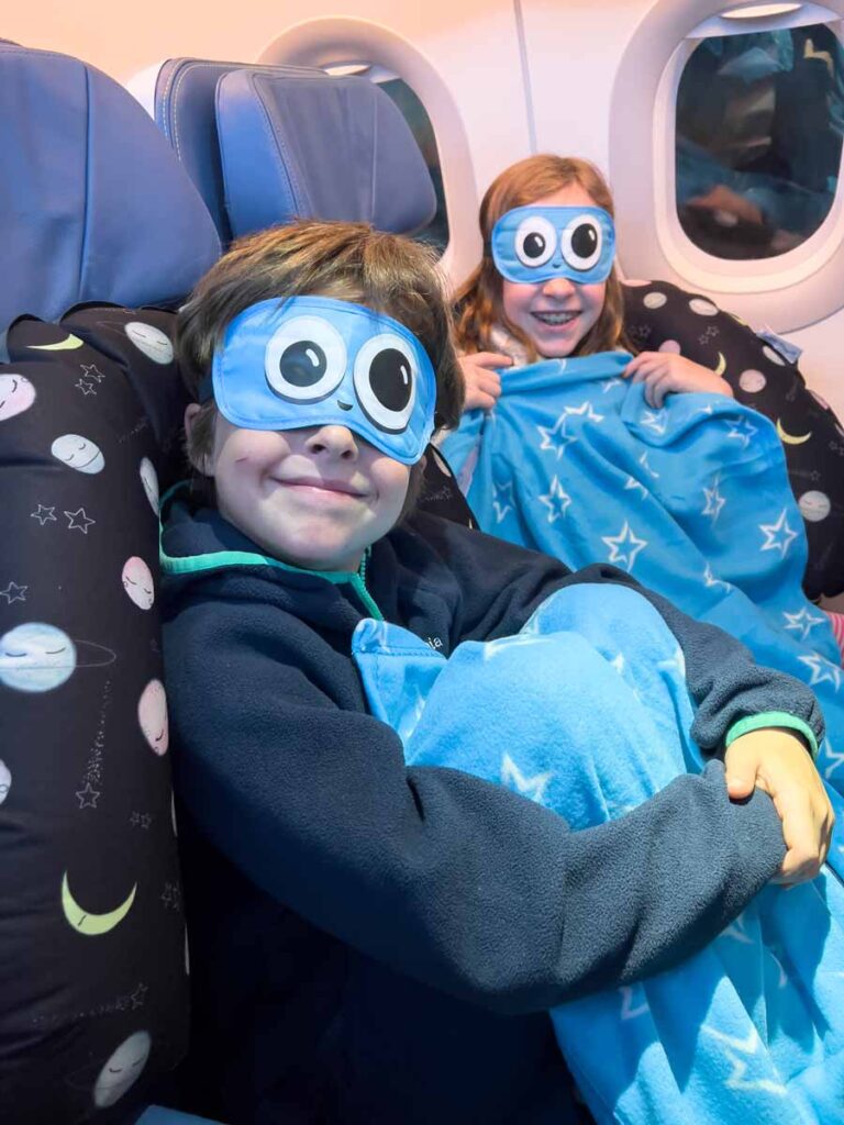 Two kids wearing funny eyemasks are ready to sleep on an airplane using their Seat to Sleep travel pillow for kids.