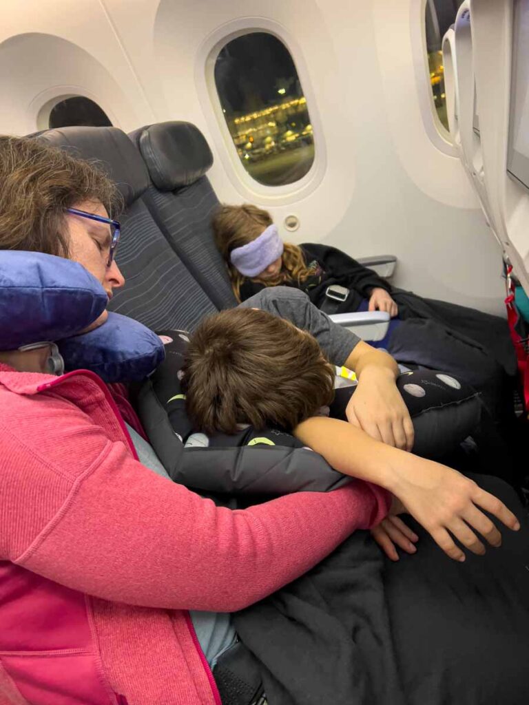 Celine Brewer, owner of BabyCanTravel.com, is asleep with her kids on an overnight flight. Both ids are using Seat to Sleep airplane pillows for kids.